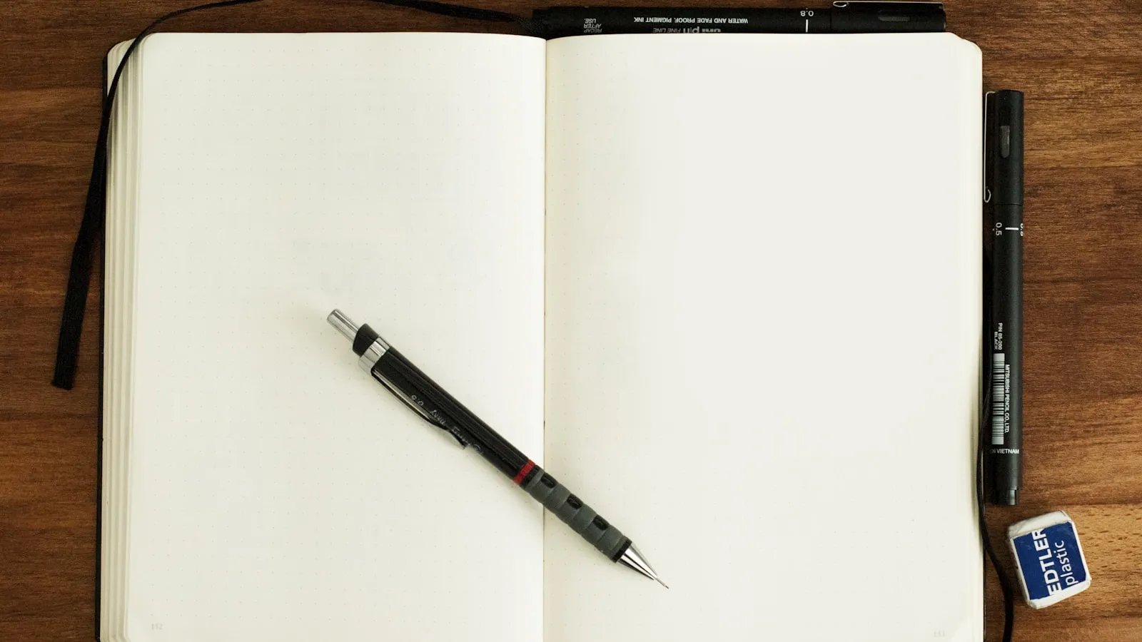 Een opengeslagen leeg notitieboek met een pen erop, op een houten tafel, met een fineliner en een gum ernaast.