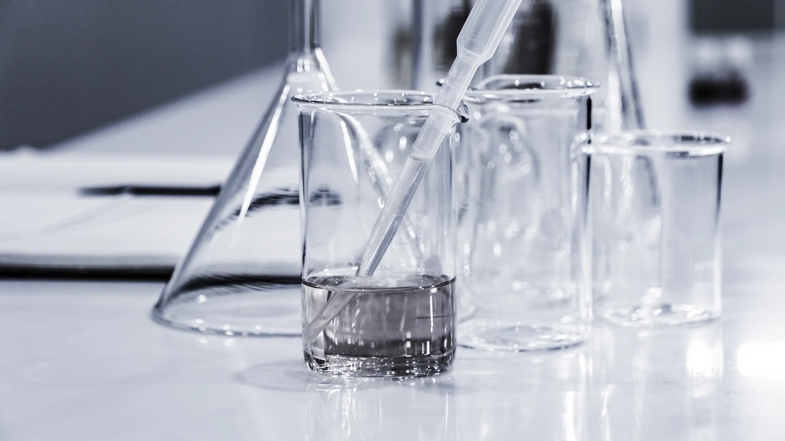 Glaswerk in een laboratorium: bekerglazen en een erlenmeyer met een pipet op een witte werkbank.