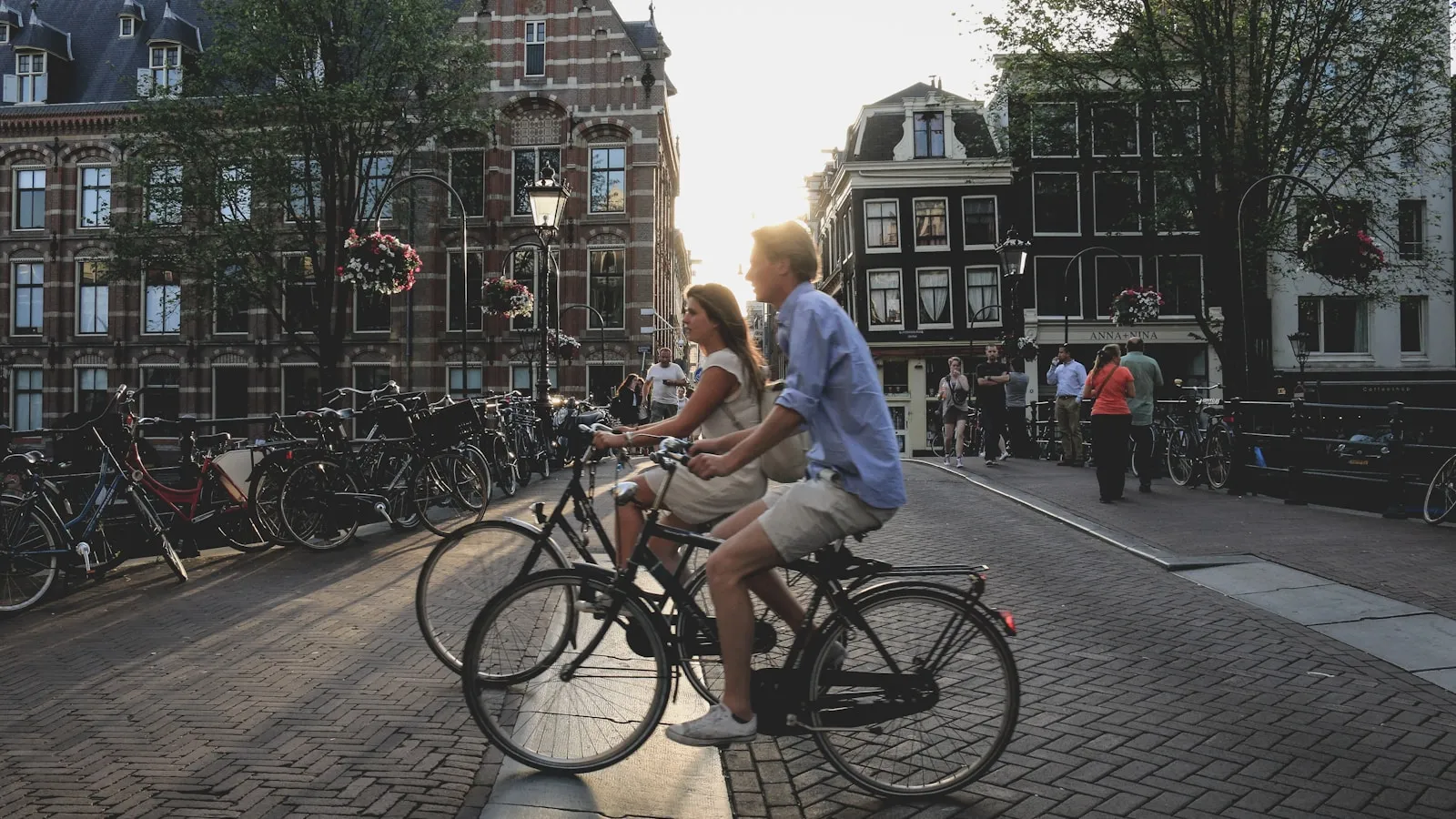 Een man en een vrouw fietsen in de ochtendzon over een Amsterdamse gracht, langs geparkeerde fietsen.
